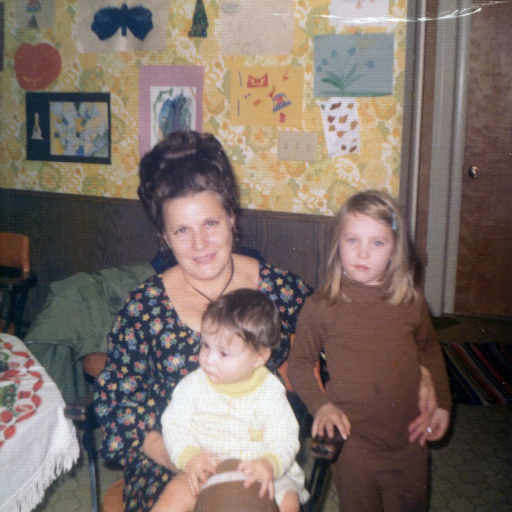 Woman with a beehive holding a baby and standing next to a child in a 70s-style living room